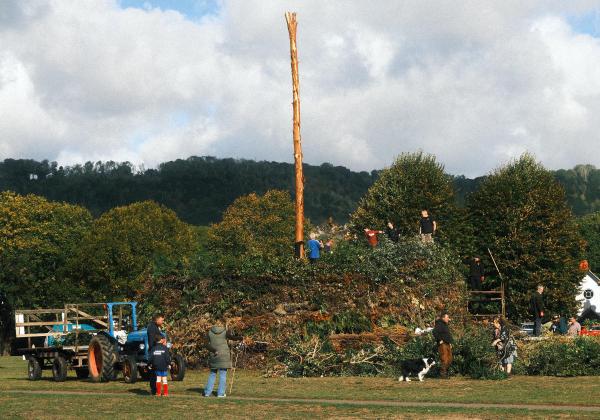 A group of people gathered in a field around a large freshly cut tree trunk, with a blue tractor and trailer standing by under a cloudy sky.